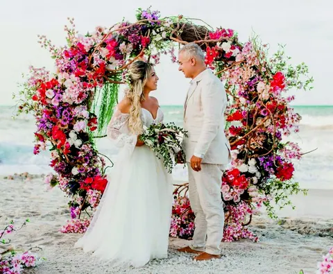 Noiva e noivo de mãos dadas diante de um grande arco de flores coloridas na praia