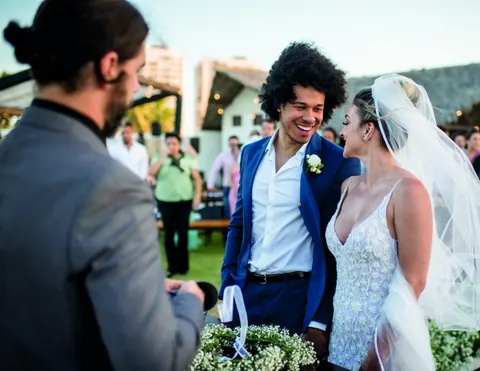 Celebrante de costas e noivos sorrindo de mãos dadas, com o véu ao vento, em cerimônia ao ar livre